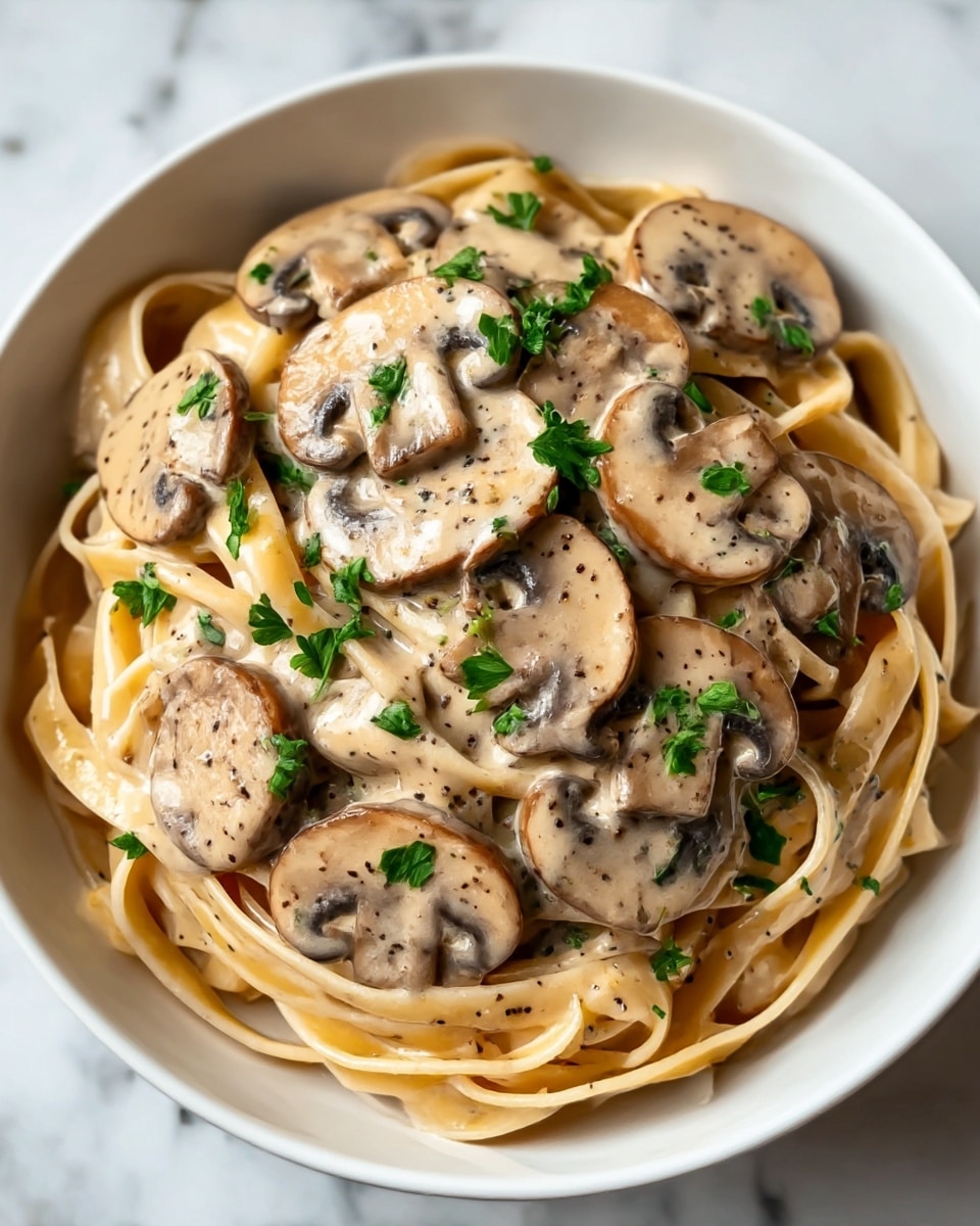 A white bowl is filled with creamy fettuccine pasta, covered in a light beige sauce with visible specks of black pepper. On top, there are many cooked mushroom slices that are brown with soft edges, also coated in the sauce. Small pieces of fresh green parsley are scattered over the mushrooms and pasta. The thick sauce gives the dish a smooth, rich look, and the pasta strands are twisted neatly below the mushrooms. The bowl is placed on a white marbled surface. Photo taken with an iphone --ar 4:5 --v 7