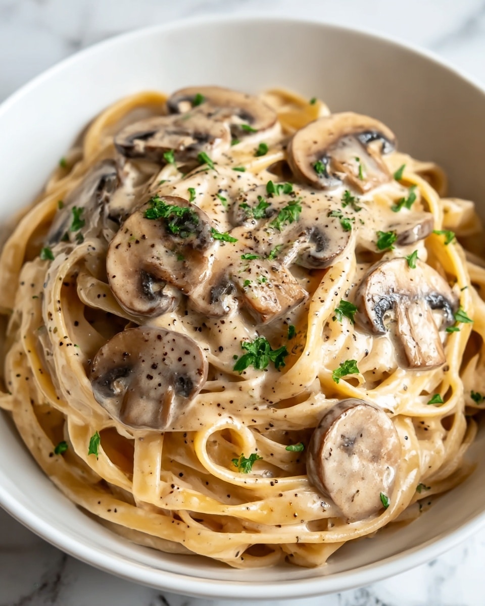 A white bowl filled with a creamy mushroom pasta dish, showing one main layer of thick fettuccine noodles mixed in a light beige cream sauce dotted with black pepper. On top, sliced brown mushrooms are coated in the same creamy sauce, evenly spread across the noodles with small pieces of bright green parsley sprinkled over the dish for color contrast. The bowl sits on a white marbled texture, and the pasta looks rich and glossy with a soft texture. photo taken with an iphone --ar 4:5 --v 7