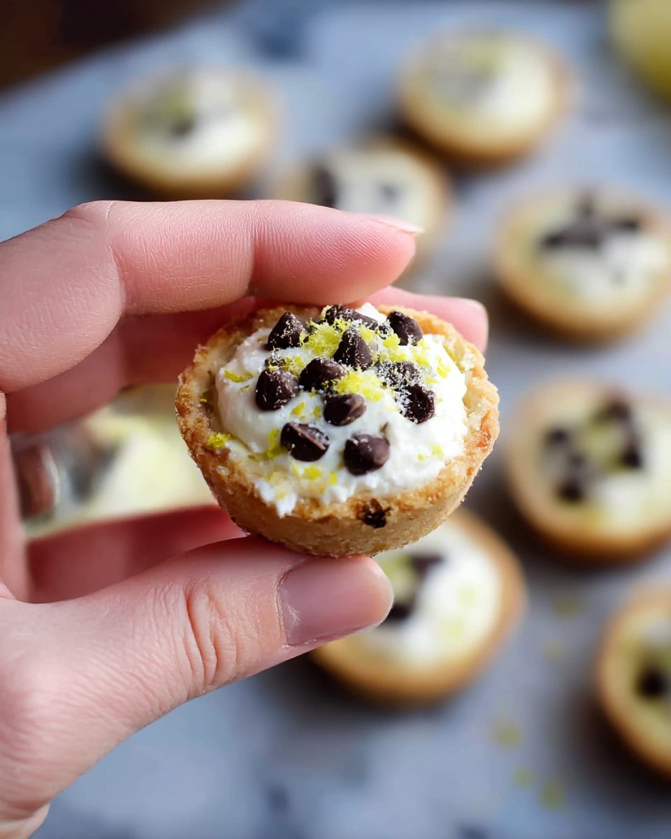 A close-up view of a small tart held by a woman's hand shows three main layers: a golden brown crispy tart shell at the bottom, a thick, creamy white filling in the middle, and dark brown chocolate chips scattered on top, with a light dusting of yellow-green zest adding color contrast. In the blurred background, several identical tarts are arranged on a white marbled surface, each decorated with similar layers and toppings, creating a cozy, inviting scene. photo taken with an iphone --ar 4:5 --v 7