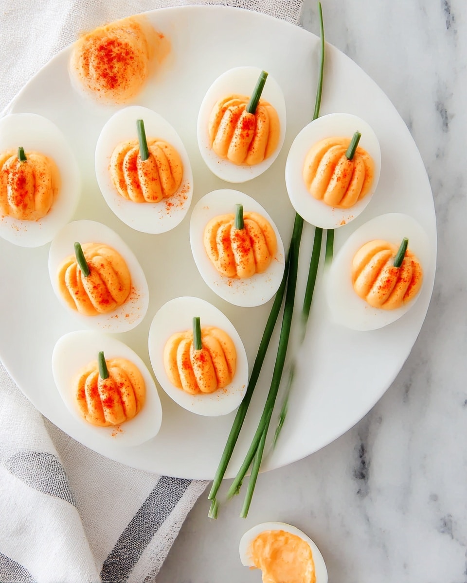 A white plate holds eight deviled eggs cut in half, each with a smooth white egg white layer and a creamy, orange filling shaped like a small pumpkin with ridges on top. Each filling has a sprinkle of red powder, likely paprika, for an added touch of color and a short green chive piece inserted at the top center to resemble a pumpkin stem. Two long green chives rest on the plate beneath the eggs. Below the plate on a white marbled surface, one deviled egg half shows a bite taken out, revealing the soft, smooth orange filling inside. The plate sits partly on a folded white and grey cloth. Photo taken with an iphone --ar 4:5 --v 7