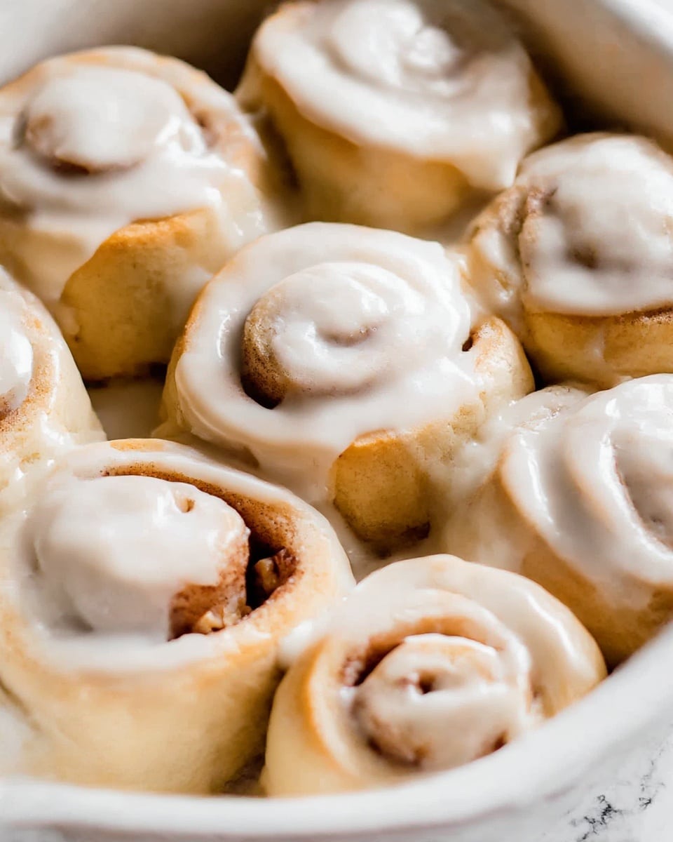 A close-up view of nine cinnamon rolls placed closely in a white baking dish, each roll showing a soft, golden-brown dough base with visible layers spiraled with light brown cinnamon filling; a thick layer of creamy white icing is spread unevenly on top, dripping slightly down the sides, making the texture look soft and smooth; the rolls are arranged in a grid pattern with some parts of the cinnamon filling peeking through the icing. The surface below is a white marbled texture. photo taken with an iphone --ar 4:5 --v 7