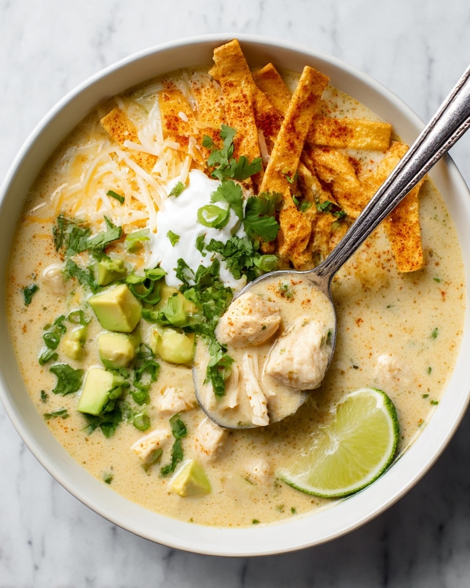 A deep bowl filled with creamy, pale yellow-white soup that has chunks of chicken and small bits of green herbs mixed throughout, topped with a few layers: crispy tortilla strips that are golden brown and dusted with spices on one side, a mix of shredded white and orange cheese scattered over and near the tortilla strips, small cubes of light green avocado spread around, chopped bright green scallions sprinkled evenly, a dollop of white sour cream near the center with fresh green cilantro leaves on top, and a thin, vibrant lime wedge resting against the edge of the bowl. A silver spoon holds a scoop of the creamy soup with chicken pieces, resting inside the bowl. The bowl is white and sits on a white marbled textured surface. Photo taken with an iphone --ar 4:5 --v 7