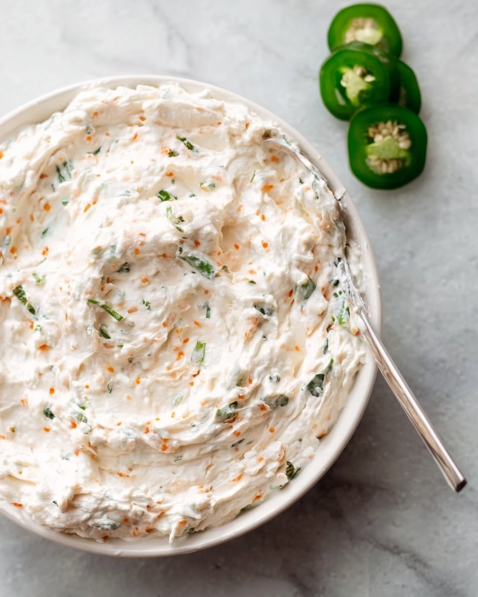 The image shows a white bowl filled with a creamy, thick dip that has a slightly whipped texture. The dip is mostly white with small bits of green herbs and tiny orange flecks mixed throughout. A silver spoon is partially immersed in the dip on the right side of the bowl. Next to the bowl on the white marbled surface, there are two slices of fresh green jalapeño pepper. The overall look is fresh and inviting, with the dip having a smooth yet slightly chunky consistency. photo taken with an iphone --ar 4:5 --v 7