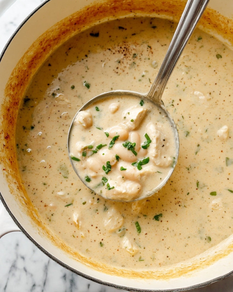 The image shows a large white pot filled with a creamy soup that has a light beige color with visible small green herbs and soft white beans throughout. There are chunks of tender, white chicken pieces mixed evenly in the soup. A silver ladle is partially dipped into the soup, resting against the side of the pot and holding a scoop of the thick mixture, with some chopped green herbs sprinkled on top. The inside edge of the pot is slightly stained with the creamy soup, showing specks of black pepper and spices. The pot is placed on a white marbled surface. photo taken with an iphone --ar 4:5 --v 7