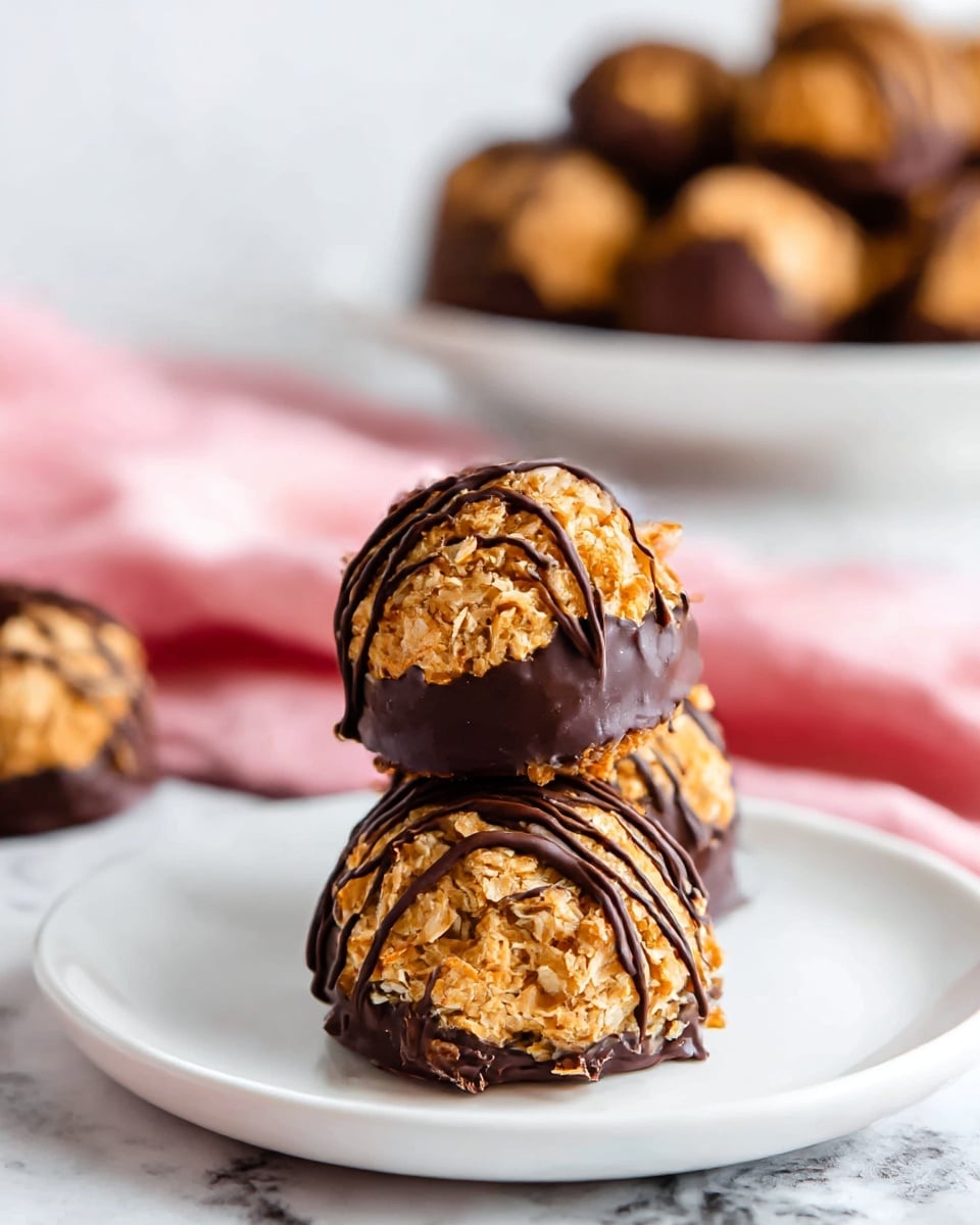 A close-up view shows three round cookie balls stacked on a white plate, each with a crunchy, crumbly golden-brown texture. The bottom of each cookie is dipped in smooth dark chocolate, creating a shiny dark base, while thin lines of dark chocolate drizzle decorate the top and sides, adding a glossy contrast to the golden cookie surface. In the background, there is a white plate filled with more cookie balls, all set on a white marbled surface with a soft pink cloth partially visible. Photo taken with an iphone --ar 4:5 --v 7
