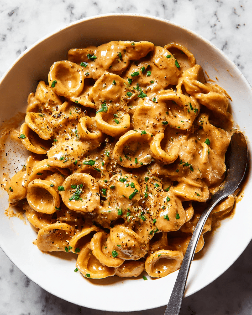A white bowl filled with about three layers of orecchiette pasta covered in a thick, creamy brown sauce. The pasta is small, round, and slightly curved, with the sauce clinging to each piece, giving a glossy texture. The dish is topped with chopped green herbs scattered evenly and cracks of black pepper adding specks of texture. A spoon rests partially submerged in the pasta on the right side of the bowl. The background is a white marbled texture surface which contrasts softly with the warm tones of the pasta and sauce. photo taken with an iphone --ar 4:5 --v 7