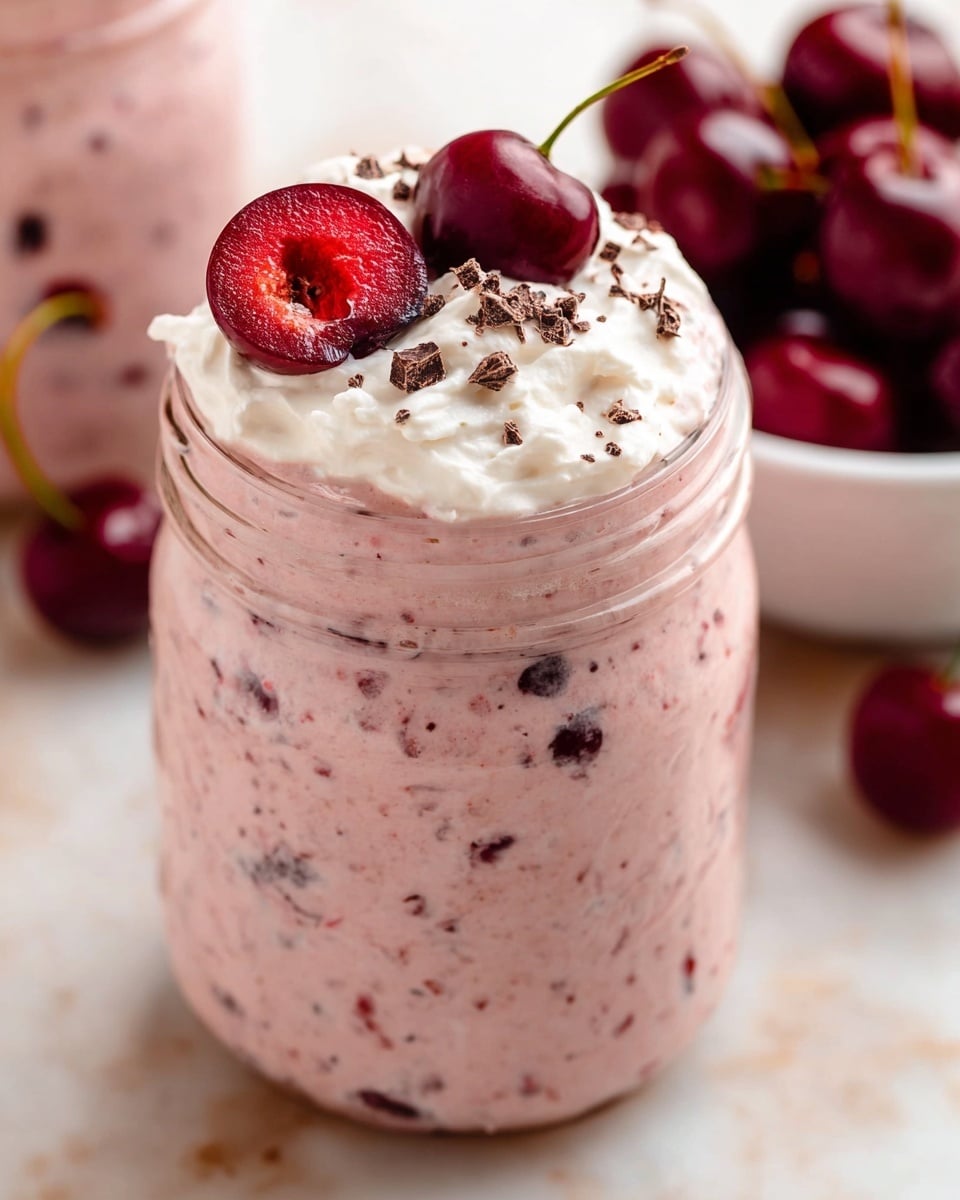 The image shows a clear glass jar filled with a thick, light pink creamy mixture that has small chunks of dark red cherries inside, giving it a speckled texture. On top of the jar, there is a dollop of white whipped cream, sprinkled with small dark brown chocolate pieces. Three whole cherries with stems, one cut in half revealing its bright red interior and seed, rest on the whipped cream. The jar is placed on a white marbled surface, and a white bowl filled with whole cherries is blurry in the background. Photo taken with an iphone --ar 4:5 --v 7