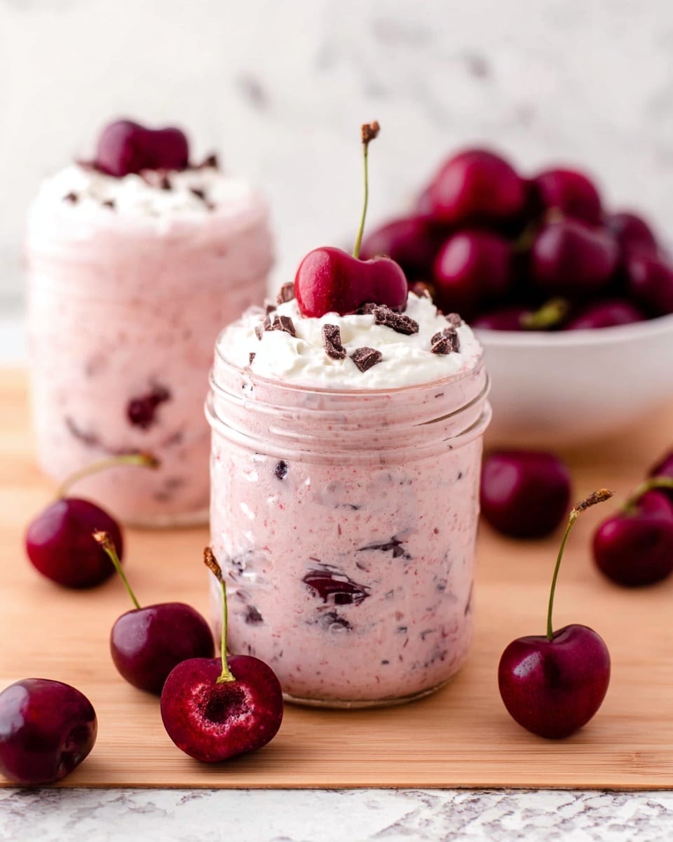 The image shows two clear glass jars filled with a thick cherry yogurt mix that is light pink with visible dark cherry pieces scattered inside. Each jar has a dollop of white cream on top, sprinkled with small dark chocolate bits. Three whole cherries, one cut in half showing its red inside and pit, sit atop the cream on the front jar. Around the jars, there are several whole cherries placed on a light wooden surface, and a white bowl filled with more fresh cherries is in the background. The overall scene sits against a white marbled texture. photo taken with an iphone --ar 4:5 --v 7