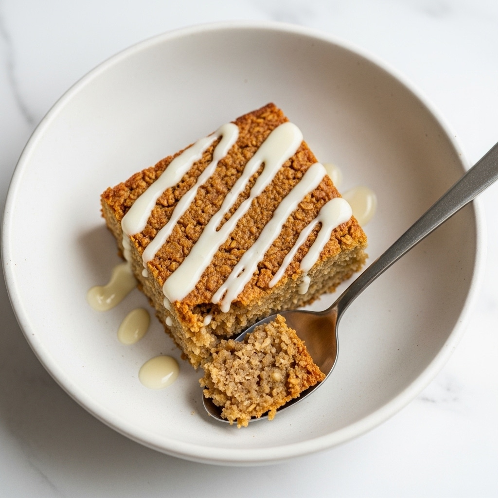 A single square piece of baked oatmeal with a rough, golden-brown texture sits in a white bowl with a matte, slightly rough surface. The oatmeal bar is topped with light white cream sauce drizzled unevenly across the top. A small portion is scooped with a silver spoon resting in the bowl, showing the moist, chunky inside of the oatmeal. The scene is set on a white marbled texture. photo taken with an iphone --ar 4:5 --v 7