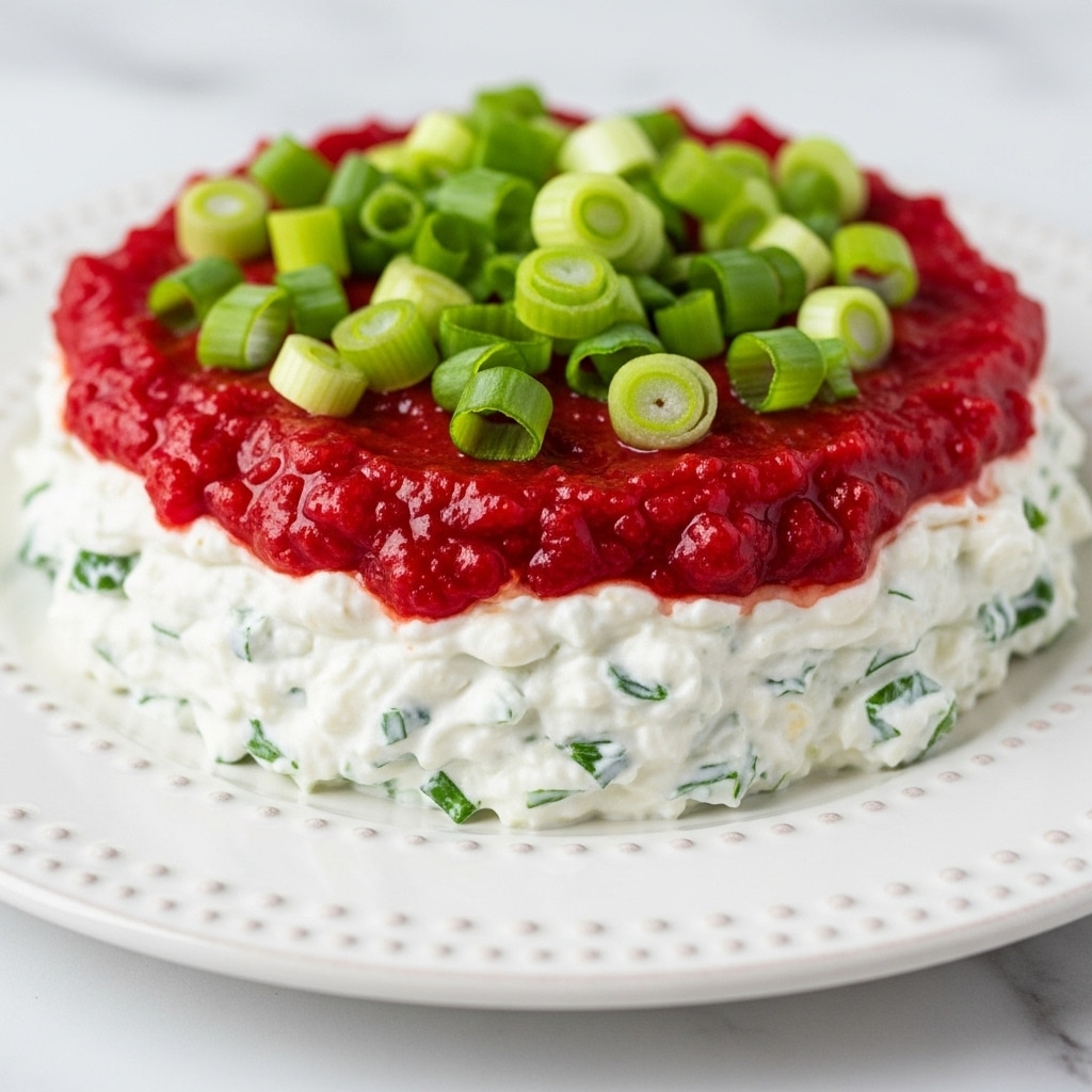 A close-up of a three-layer dip on a white plate with a textured edge, sitting on a white marbled surface. The bottom layer is a creamy white spread with small bits of green herbs mixed in, spread evenly close to the plate’s edge. The middle layer is a thick, bright red sauce with a glossy texture, covering most of the cream layer. The top layer features scattered slices of bright green scallions, adding a fresh contrast to the red sauce. Photo taken with an iphone --ar 4:5 --v 7