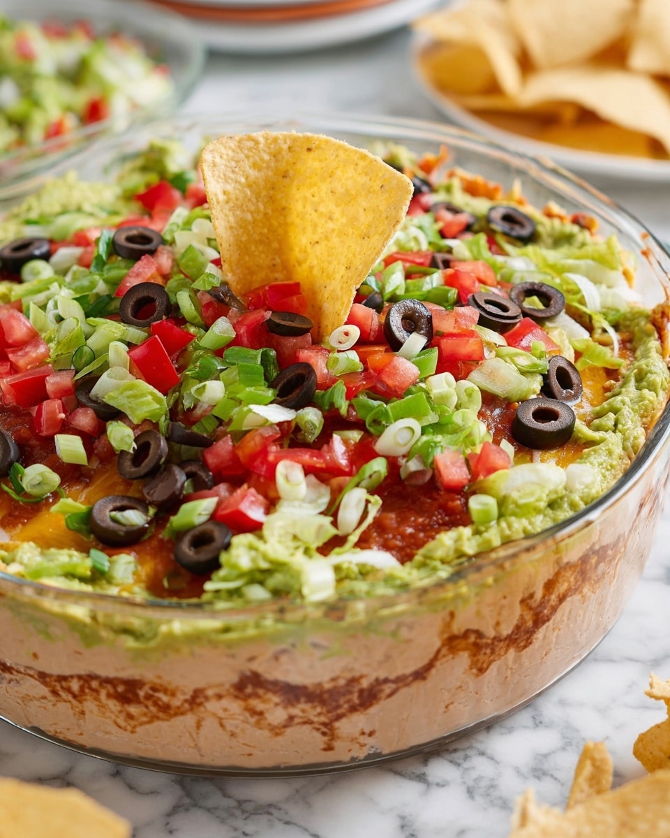 The image shows a clear glass dish filled with a layered dip. The bottom layer is beige refried beans with a smooth texture, followed by a layer of melted cheese mixed with some salsa that is orange-red in color. Above this, there is a green guacamole layer, topped with a mix of chopped fresh ingredients including bright green lettuce, red diced tomatoes, sliced black olives, and green onion rings that cover the top generously. A single beige tortilla chip is inserted vertically near the center of the dish. The dish is set on a white marbled surface, with blurred plates and more tortilla chips in the background. photo taken with an iphone --ar 4:5 --v 7