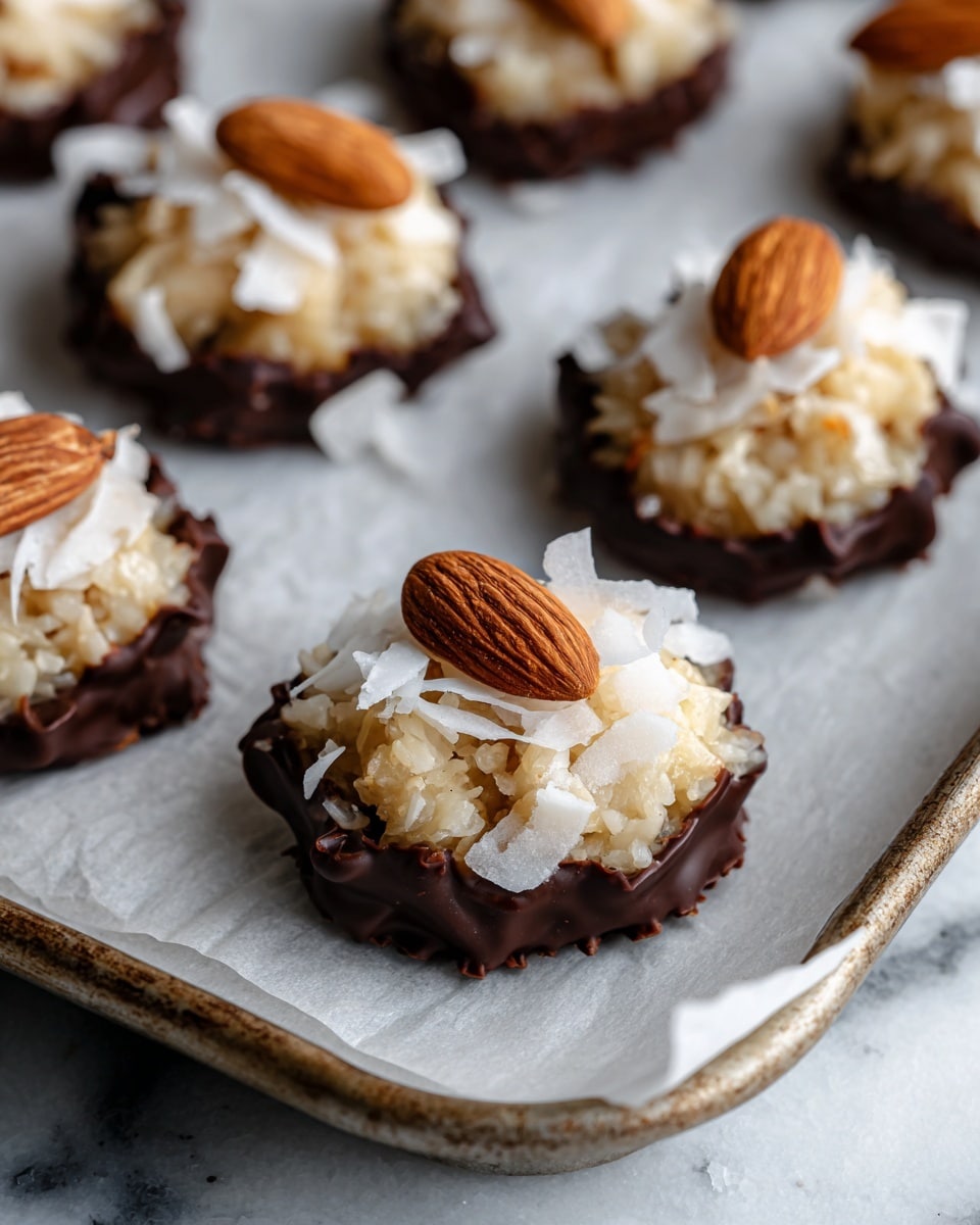 Small round treats are placed on white parchment paper over a baking tray. Each treat has three visible layers: the bottom layer is rough and coated in dark brown chocolate, the middle layer is a chunky pale beige coconut mix with larger white shredded coconut pieces, and the top layer shows one or two light brown whole almonds placed as decoration. The edges of the chocolate base are uneven, and the coconut mixture is piled in a small mound above it. The background surface has a white marbled texture. photo taken with an iphone --ar 4:5 --v 7