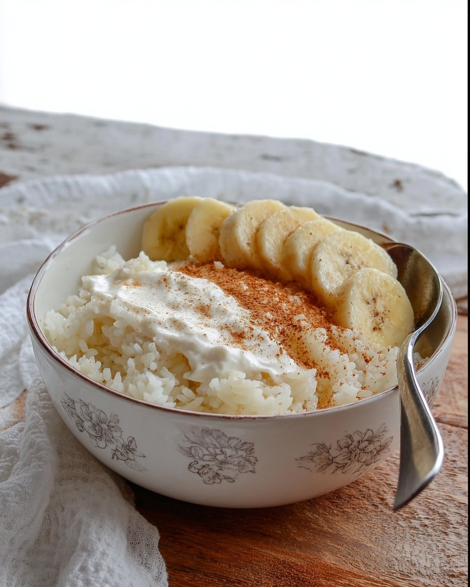 A bowl filled with three main layers: the bottom layer is soft cooked white rice with a light sprinkling of cinnamon powder, the middle layer includes creamy white yogurt spread over the rice on one side, and the top layer has light yellow banana slices placed neatly with a dusting of cinnamon. A silver spoon rests inside the bowl on the right side. The bowl itself is white with a subtle, light gray floral pattern on the inside. The bowl sits on a wooden table with a white marbled texture in the background and a piece of white cloth nearby. photo taken with an iphone --ar 4:5 --v 7