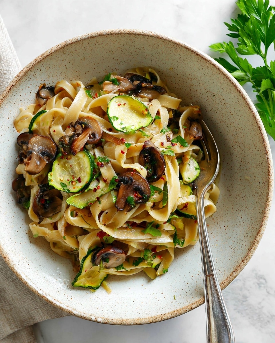 A bowl holds a serving of pasta with three visible layers of ingredients. The base layer is light cream-colored flat noodles twisted and intertwined with a slightly shiny, soft texture. Spread throughout the noodles are dark brown sautéed mushrooms that look tender with a slight gloss. Slices of cooked green zucchini with a soft texture and some char marks are mixed evenly with the pasta and mushrooms. Small bits of herbs and red pepper flakes are scattered lightly across the dish, adding tiny dots of green and red color. A sprig of fresh green parsley rests on the side for garnish. A silver fork leans on the side of the bowl. The bowl itself is white with a slightly rough, speckled texture and is set on a white marbled surface. Photo taken with an iphone --ar 4:5 --v 7