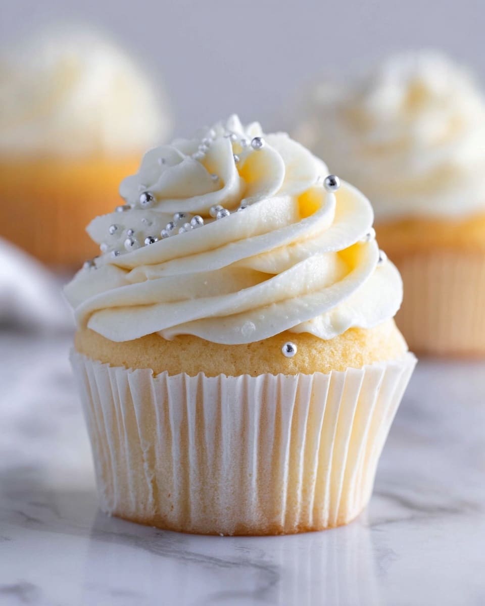A single cupcake with two layers is shown up close on a white marbled surface. The bottom layer is a light golden sponge cake wrapped in a white paper liner with vertical pleats. On top, there is a thick swirl of creamy white frosting with a smooth, fluffy texture, decorated with small shiny silver balls scattered across its curves. The background is softly blurred, showing more cupcakes in the distance. Photo taken with an iphone --ar 4:5 --v 7