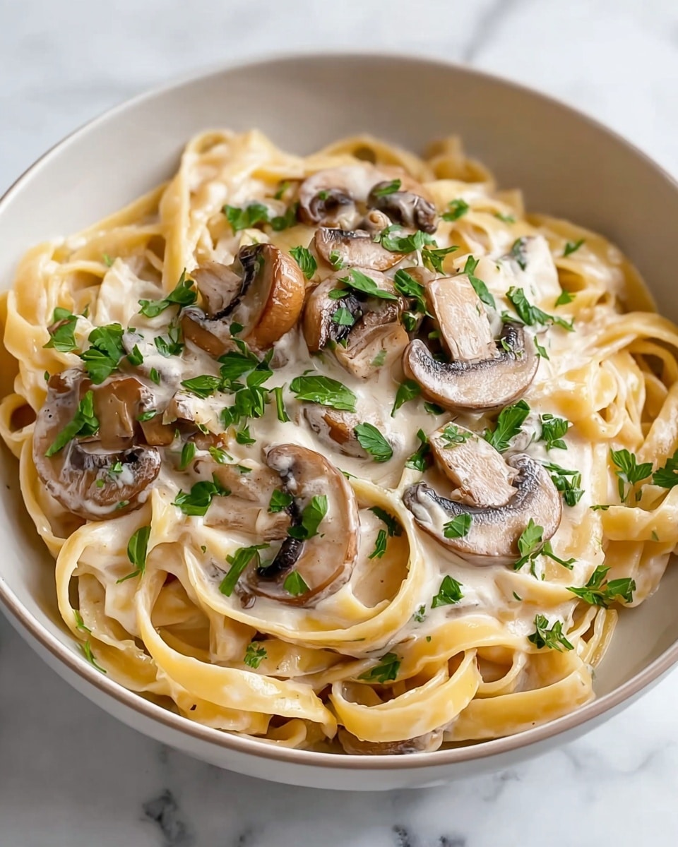 This image shows a white bowl filled with creamy fettuccine pasta. The pasta noodles are smooth and pale yellow, tangled softly in the bowl's base. On top, there is a rich layer of creamy white sauce covering the noodles. Sliced mushrooms with a light brown color and soft texture sit scattered across the surface. Fresh, chopped green parsley leaves are sprinkled over the mushrooms and pasta, adding a pop of bright color. The bowl is placed on a white marbled surface. photo taken with an iphone --ar 4:5 --v 7