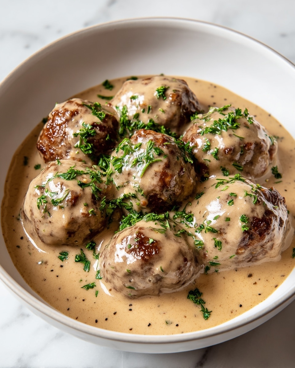 A white bowl holds seven browned meatballs that are fully covered in a thick, creamy light brown sauce. Fresh chopped green herbs are sprinkled on top, adding a touch of bright color to the dish. The sauce has a smooth texture with small specks of seasoning visible throughout. The meatballs sit close together in the bowl, partially submerged in the sauce. The bowl is placed on a white marbled surface. photo taken with an iphone --ar 4:5 --v 7