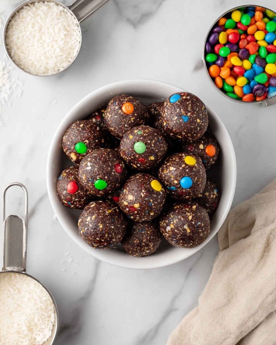 A white bowl filled with about a dozen round dark brown energy balls, each studded with small colorful candy pieces in red, yellow, green, blue, orange, and purple, showing a slightly rough texture with visible bits of nuts or seeds. Around the bowl, a metal measuring cup filled with tiny colorful candies sits at the top right. At the bottom left, another metal measuring cup holds shredded white coconut. The background is a clean white marbled surface with a beige cloth partially visible in the lower right corner. Photo taken with an iphone --ar 4:5 --v 7