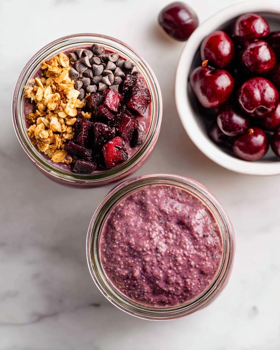 The image shows two glass jars filled with a thick, purple chia pudding. One jar is topped with three distinct sections: golden brown granola on the left, dark chocolate chips in the middle, and deep red cherry pieces on the right, all placed on top of the purple pudding layer. The other jar is filled all the way with just the purple pudding, showing its smooth, textured surface. To the right, there is a white bowl filled with whole dark red cherries. The setup is on a white marbled surface, giving a clean and fresh look. photo taken with an iphone --ar 4:5 --v 7