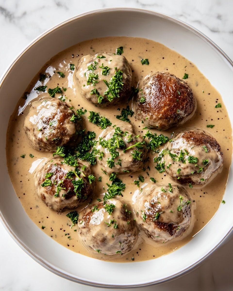 A white bowl with a thin gray rim holds seven browned meatballs that are completely covered with light brown creamy sauce. The sauce has a smooth texture with small specks of seasoning. Bright green chopped parsley is sprinkled on top, adding a fresh look and color contrast. The bowl rests on a white marbled surface that highlights the dish's warm and rich appearance. photo taken with an iphone --ar 4:5 --v 7